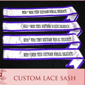 May include: Four white satin sashes with purple trim and the text "MISS * MISS TEEN VIETNAM NORCAL DELEGATE" in black. The sashes are arranged diagonally against a black background. The bottom of the image reads "CUSTOM LACE SASH".