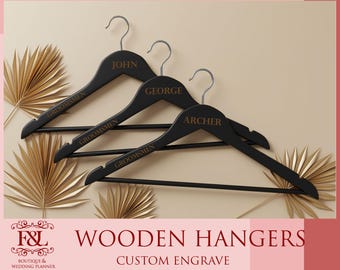 May include: Three black wooden hangers with silver hooks, each engraved with a name in gold lettering: John, George, and Archer. The word "Groomsmen" is also engraved on each hanger. The image includes gold-colored decorative leaves and the text "Wooden Hangers, Custom Engrave".