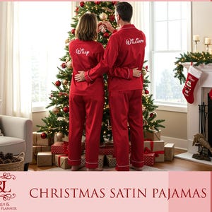 May include: Two people wearing matching red satin Christmas pajamas, standing in front of a decorated Christmas tree. The pajamas have white trim and personalized names on the back. The image also includes a fireplace with stockings and wrapped gifts.