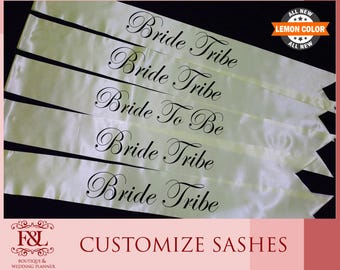 May include: Cream-colored satin sashes with black script that reads "Bride Tribe" and "Bride To Be". The sashes are arranged in a fan shape. The image also includes text that says "Lemon Color" and "Customize Sashes".