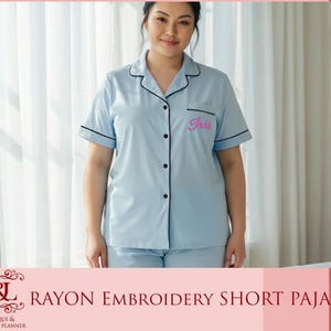 May include: Light blue rayon short pajama set with black trim. The pajama top has short sleeves, a button-down front, and the name "Iris" embroidered in pink. The set includes matching shorts. The image also includes the text "RAYON EMBROIDERY SHORT PAJAMA".