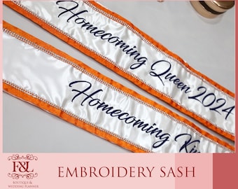 May include: Two white satin sashes with orange trim and rhinestone accents. The sashes have the words "Homecoming Queen 2024" and "Homecoming King" embroidered in navy blue script. The sashes are for a homecoming event.
