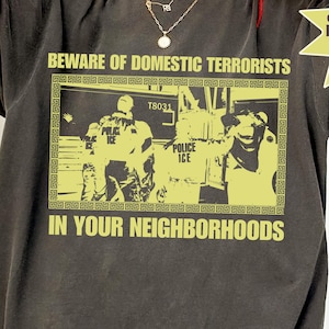 May include: Dark gray t-shirt with a yellow graphic that reads "BEWARE OF DOMESTIC TERRORISTS" above an image of police officers. The text "IN YOUR NEIGHBORHOODS" is below the image. The shirt also has a logo that says "Profits donated to Immigrant Defense Project."