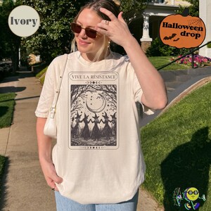 A model wearing an ivory oversized T-shirt featuring a black tarot card-style design with four witches holding hands under a crescent moon and stars. The top of the card reads "VIVE LA RÉSISTANCE" with moon phase symbols above and below the illustration. The model also wears sunglasses and carries a white shoulder bag. A pumpkin graphic labeled "Halloween drop" is in the top right corner.