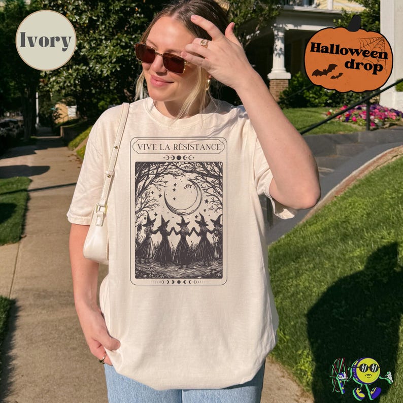 A model wearing an ivory oversized T-shirt featuring a black tarot card-style design with four witches holding hands under a crescent moon and stars. The top of the card reads "VIVE LA RÉSISTANCE" with moon phase symbols above and below the illustration. The model also wears sunglasses and carries a white shoulder bag. A pumpkin graphic labeled "Halloween drop" is in the top right corner.