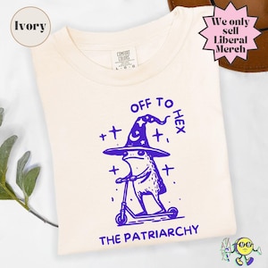 May include: An ivory t-shirt featuring a purple graphic of a frog wearing a witch hat and riding a scooter. The text above the frog reads OFF TO HEX and the text below reads THE PATRIARCHY. The shirt has a Comfort Colors label at the neck.