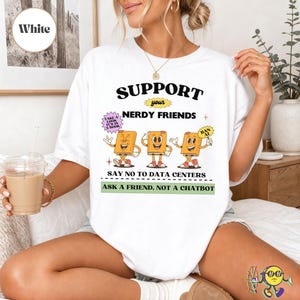 Könnte beinhalten: Ein weißes T-Shirt mit dem Text SUPPORT YOUR NERDY FRIENDS, SAY NO TO DATA CENTERS und ASK A FRIEND, NOT A CHATBOT. Die Grafik zeigt drei gezeichnete Bücher mit Armen und Beinen sowie Sprechblasen mit dem Text TAKE A LOOK IT'S IN A BOOK und BAN AI.