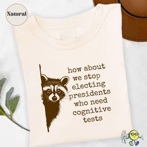 May include: Cream-colored t-shirt featuring a brown raccoon graphic peeking from behind a vertical line. Text on the shirt reads, "how about we stop electing presidents who need cognitive tests."