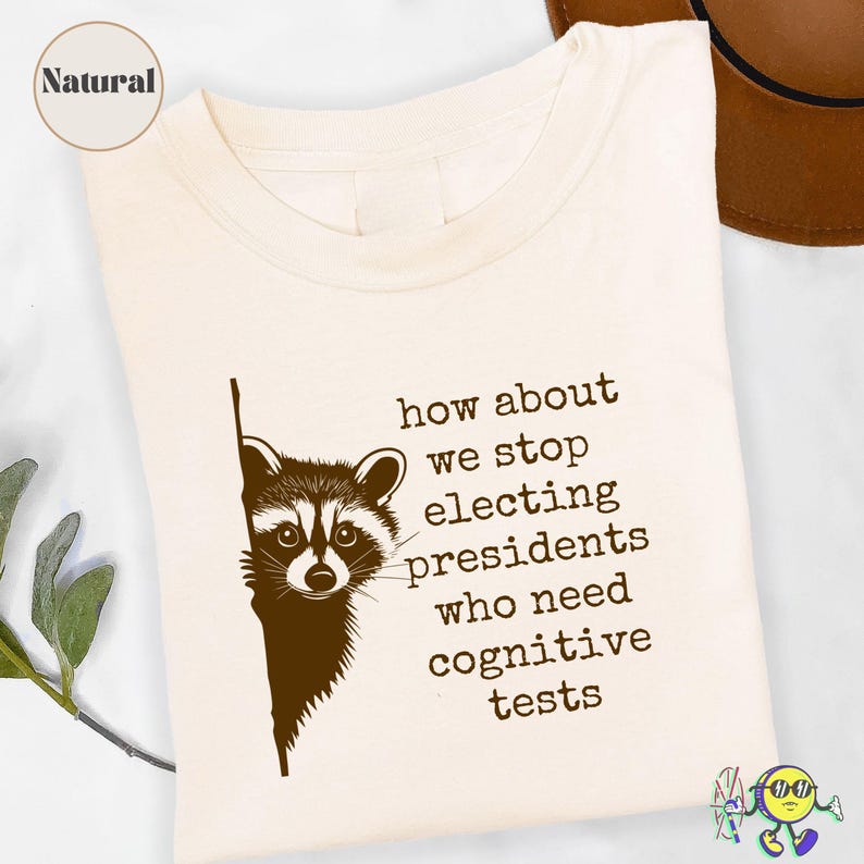 May include: Cream-colored t-shirt featuring a brown raccoon graphic peeking from behind a vertical line. Text on the shirt reads, "how about we stop electing presidents who need cognitive tests."