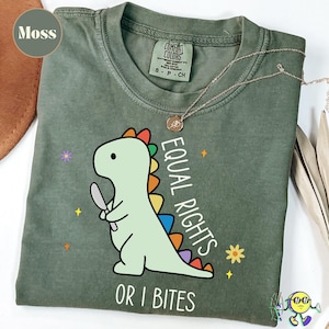 May include: Moss green t-shirt with a cartoon dinosaur holding a knife. The dinosaur has a rainbow spine and the text "EQUAL RIGHTS OR I BITES". A gold necklace with a pendant is on the shirt.