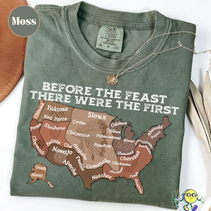 May include: A moss green t-shirt with the text "BEFORE THE FEAST THERE WERE THE FIRST" above a map of the United States, highlighting various Native American tribes. A gold necklace rests on the shirt.