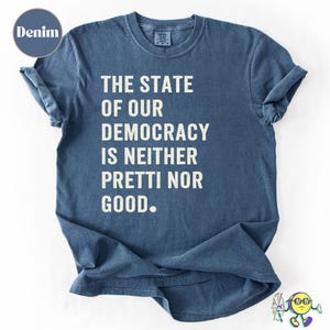 May include: A denim blue t-shirt with rolled sleeves. The white text on the shirt reads: "THE STATE OF OUR DEMOCRACY IS NEITHER PRETTI NOR GOOD." The t-shirt is made of a soft material and is suitable for casual wear.