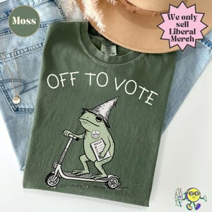Puede incluir: Una camiseta verde musgo con el gráfico de una rana con sombrero puntiagudo, montada en un patinete y sosteniendo una papeleta. El texto "OFF TO VOTE" aparece arriba y una pegatina "I VOTED" se ve en el pecho de la rana.