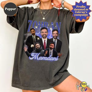 A person wearing an oversized, faded black "Pepper" color T-shirt featuring a collage of five photos of a man in different poses and suits. The text "ZOHRAN" in large blue letters with sparkles and lightning in the background is above the images, and "Mamdani" in cursive blue font is below. The design has a vintage, retro poster style. Profits from this purchase are donated to Zohran's campaign.