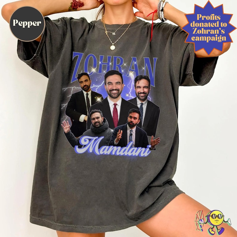A person wearing an oversized, faded black "Pepper" color T-shirt featuring a collage of five photos of a man in different poses and suits. The text "ZOHRAN" in large blue letters with sparkles and lightning in the background is above the images, and "Mamdani" in cursive blue font is below. The design has a vintage, retro poster style. Profits from this purchase are donated to Zohran's campaign.