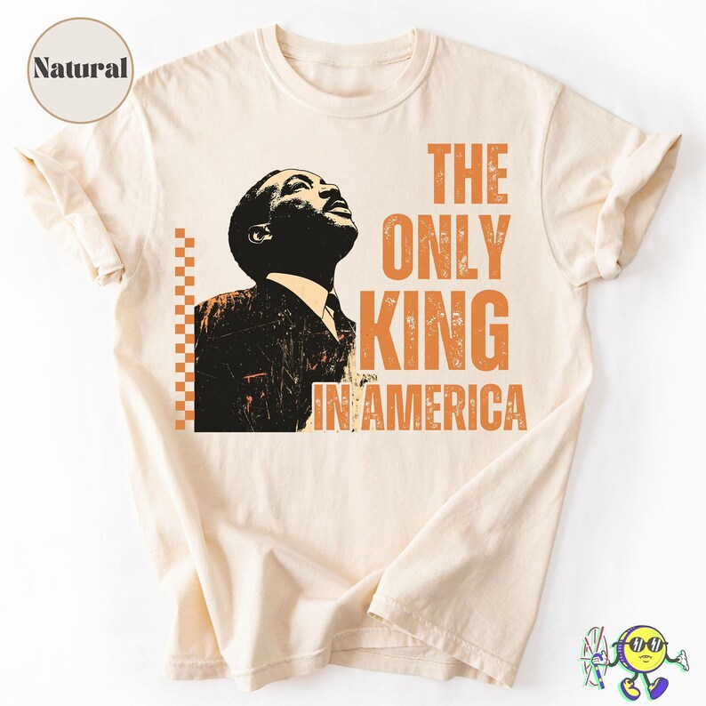 Natural-colored short-sleeve T-shirt with a graphic print of a man in a suit looking upward in black and beige. To the right of the image, bold orange text reads "THE ONLY KING IN AMERICA." An orange vertical checkerboard pattern is placed to the left of the man’s image. The sleeves are casually rolled up. A circular label with the word "Natural" is visible in the upper left corner.