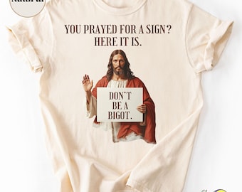 Può includere: T-shirt color crema con la scritta "YOU PRAYED FOR A SIGN? HERE IT IS." Sotto, un'immagine di Gesù che tiene un cartello con la scritta "DON'T BE A BIGOT."