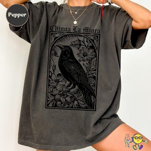 Può includere: T-shirt grigio scuro con un'illustrazione di corvo nero all'interno di una cornice floreale. Il testo "Chinga La Migra" è sopra il corvo. La maglietta ha un aspetto vintage.