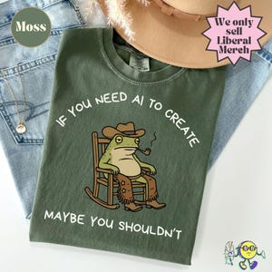 Może przedstawiać: Oliwkowozielony t-shirt z grafiką żaby w kowbojskim kapeluszu i czapsach, siedzącej w fotelu bujanym i palącej fajkę. Napis "IF YOU NEED AI TO CREATE MAYBE YOU SHOULDN'T" jest umieszczony białymi literami wokół ilustracji na koszulce.
