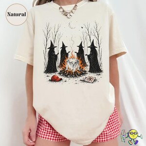 May include: A natural-colored t-shirt featuring a graphic illustration of four figures in black cloaks and pointed hats standing around a campfire. The scene includes a red cap and a square sign with a crossed-out symbol on the ground.