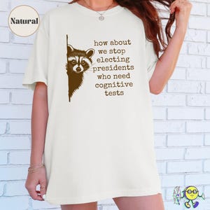 A model wearing a natural-colored oversized t-shirt featuring a dark brown graphic of a raccoon peeking from the left edge. To the right of the raccoon, text reads in all lowercase typewriter-style font: "how about we stop electing presidents who need cognitive tests." A circular label in the top left corner says "Natural."