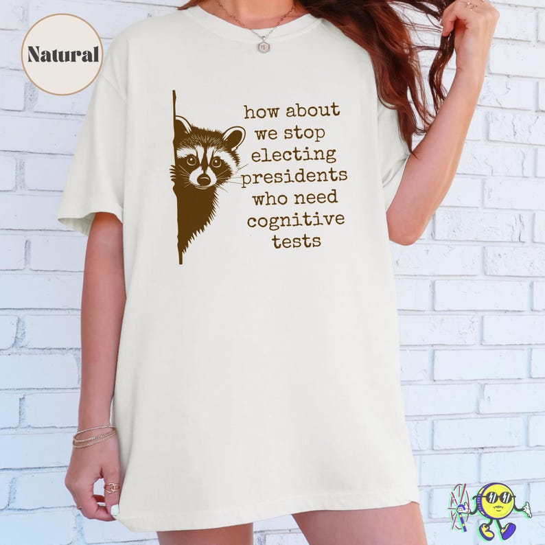 A model wearing a natural-colored oversized t-shirt featuring a dark brown graphic of a raccoon peeking from the left edge. To the right of the raccoon, text reads in all lowercase typewriter-style font: "how about we stop electing presidents who need cognitive tests." A circular label in the top left corner says "Natural."