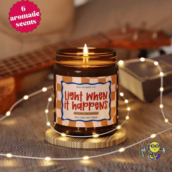May include: A brown glass jar candle with a lit wick, sitting on a circular wooden coaster. The label features an orange and white checkered border with the text "Light WHEN it happens" and "NATURAL SOY CANDLE" in red and brown lettering.