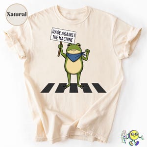 Puede incluir: Camiseta color crema con una rana de dibujos animados en un paso de cebra, sosteniendo un cartel que dice "Rage Against The Machine". La rana lleva un pañuelo azul. La camiseta tiene una etiqueta "Natural".