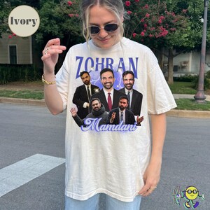 Ivory oversized T-shirt with a collage of a man in five different poses wearing suits and casual clothes. "ZOHRAN" is printed above in large purple letters with sparkles and lightning. Below the collage, "Mamdani" is written in a large cursive purple font. The fabric has a subtle texture, and the shirt has short sleeves and a crew neckline. A person wearing sunglasses models the shirt outdoors.