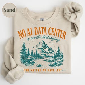 May include: A sand-colored crewneck sweatshirt featuring a graphic print with the text NO AI DATA CENTER is worth destroying and THE NATURE WE HAVE LEFT. The design includes a teal mountain landscape with pine trees and a river.