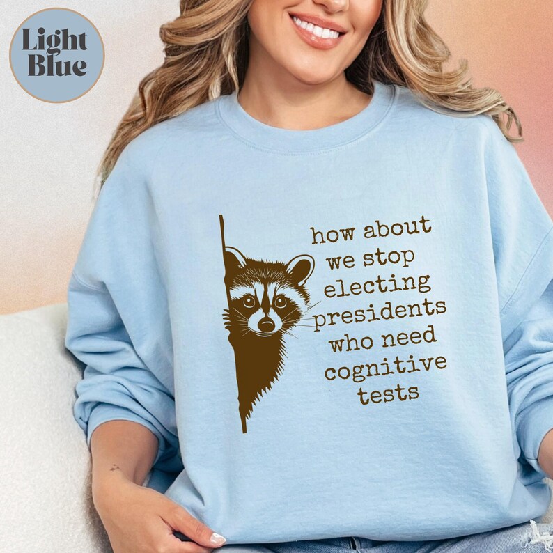 Light blue sweatshirt worn by a model, featuring a dark brown raccoon peeking from the left edge. Text in typewriter font reads, "how about we stop electing presidents who need cognitive tests," arranged in a staggered block to the right of the raccoon. The design contrasts sharply against the soft, smooth light blue fabric. A small circle with "Light Blue" text appears in the top left corner.