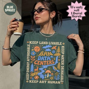 May include: A dark green t-shirt featuring a colorful graphic print with the text "KEEP LAND LIVABLE," "BAN DATA CENTERS," "KEEP ENERGY AFFORDABLE," "KEEP WATER DRINKABLE," and "KEEP ART HUMAN" surrounded by illustrations of a sun, trees, and animals.