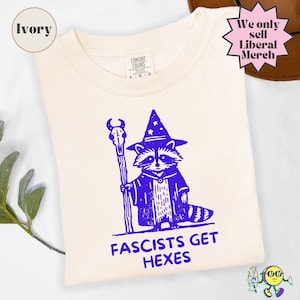 May include: An ivory t-shirt featuring a purple graphic of a raccoon wearing a wizard hat and robe, holding a staff topped with a skull. Below the raccoon, the text reads FASCISTS GET HEXES. The shirt has a Comfort Colors label at the neck.