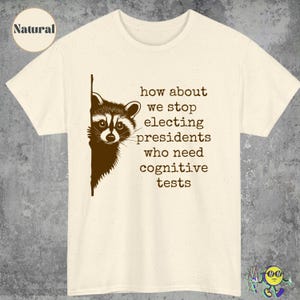 May include: Cream-colored t-shirt with a raccoon graphic and the text: "how about we stop electing presidents who need cognitive tests." The raccoon is brown and black. The shirt is made of a soft material.