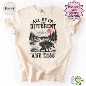 May include: An ivory t-shirt featuring a black ink graphic of a raccoon in a forest landscape with a mountain and a red sun. The text reads ALL OF US ARE DIFFERENT and NONE OF US ARE LESS. The shirt includes a circular ivory label and a pink starburst.
