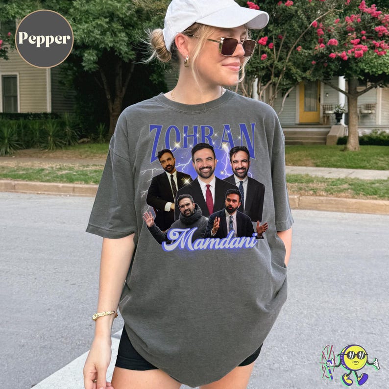 A person wears a loose, short-sleeve, Pepper-colored gray t-shirt featuring a collage of five different images of a man dressed in suits and casual wear. Above the images is the name "ZOHRAN" in bold, blue uppercase letters with white sparkles and lightning effects. Below the images, the name "Mamdani" is written in a cursive light blue font. The person pairs the shirt with black shorts, sunglasses, and a white cap.