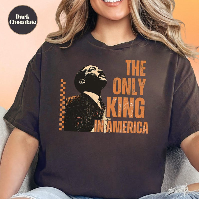 Dark chocolate-colored T-shirt featuring an orange and black graphic of a man in a suit looking upward. Next to the image, bold orange text says, "THE ONLY KING IN AMERICA." A vertical orange and black checkerboard pattern runs along the left side of the graphic. The shirt appears worn by a smiling model. A circle on the top left labels the color as “Dark Chocolate.”