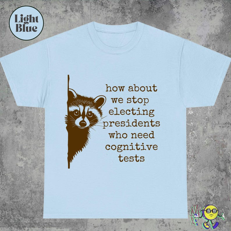 May include: Light blue t-shirt with a raccoon graphic and text that reads, "how about we stop electing presidents who need cognitive tests." The raccoon is brown and white.