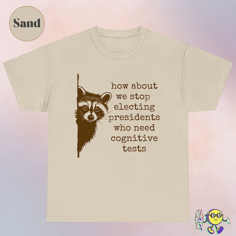 Sand-colored T-shirt featuring a brown graphic of a raccoon peeking from the left edge. To the right of the raccoon, brown typewriter-style text reads, "how about we stop electing presidents who need cognitive tests." A circle in the top left corner labels the color as "Sand."