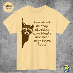 May include: A yellow haze t-shirt with a raccoon graphic peeking from the left side. The text reads, "how about we stop electing presidents who need cognitive tests."
