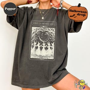 Oversized "Pepper" color tee dress featuring a vintage-style tarot card print in cream. The design shows five witches in dresses and pointed hats holding hands under a crescent moon and starry sky, framed by bare trees. Text above reads "VIVE LA RÉSISTANCE." The dress has short sleeves and a washed, slightly faded texture. Halloween drop sticker in corner.