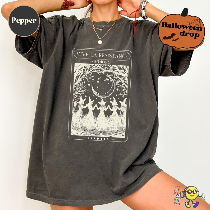 Oversized "Pepper" color tee dress featuring a vintage-style tarot card print in cream. The design shows five witches in dresses and pointed hats holding hands under a crescent moon and starry sky, framed by bare trees. Text above reads "VIVE LA RÉSISTANCE." The dress has short sleeves and a washed, slightly faded texture. Halloween drop sticker in corner.