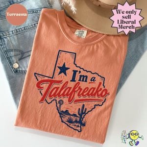 May include: A terracotta-coloured t-shirt featuring a Texas graphic with a blue star and the words "I'm a Talafreako" in red and blue. The shirt is laid on blue jeans, a necklace, and a hat.