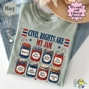 May include: A light green t-shirt with the text "CIVIL RIGHTS ARE MY JAM" and illustrations of jars labeled with various rights. The shirt also features the text "We only sell Liberal Merch".