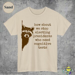 May include: Sand-colored t-shirt with a raccoon graphic peeking from the left side. The shirt features the text: "how about we stop electing presidents who need cognitive tests."