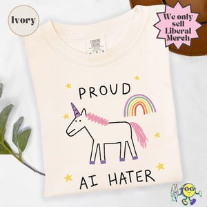 May include: An ivory t-shirt featuring a hand-drawn style illustration of a white unicorn with a pink mane and tail, a rainbow, and yellow stars. The text reads PROUD AI HATER. The shirt has a Comfort Colors label indicating a size large.