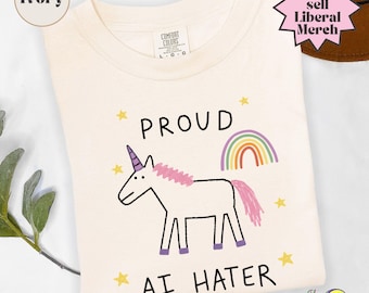 Puede incluir: Una camiseta color marfil con una ilustración estilo dibujo a mano de un unicornio blanco con crin y cola rosa, un arcoíris y estrellas amarillas. El texto dice PROUD AI HATER. La etiqueta Comfort Colors indica una talla grande.