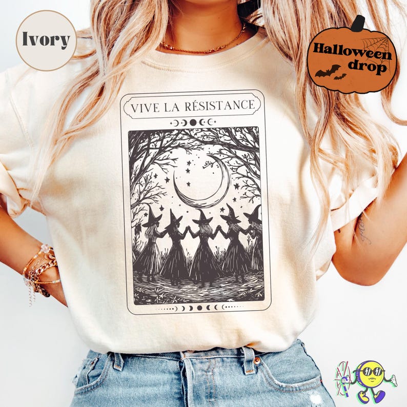 Ivory-colored t-shirt worn by a model, featuring a black graphic of five witches in pointed hats holding hands beneath a crescent moon and stars, framed by leafless trees. Above the graphic, text reads "VIVE LA RÉSISTANCE" surrounded by moon phase symbols. The design has a vintage tarot card style with mystical elements. Two overlay badges: "Ivory" in a beige circle and "Halloween drop" in an orange pumpkin shape.