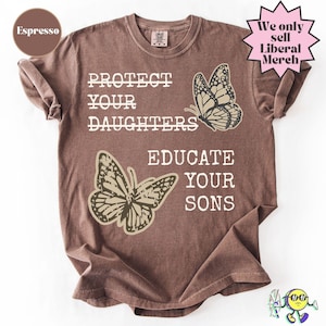 May include: An espresso-colored short-sleeve t-shirt featuring the text PROTECT YOUR DAUGHTERS and EDUCATE YOUR SONS in white lettering. Two beige butterfly illustrations are positioned next to the text. The shirt has a crew neckline.