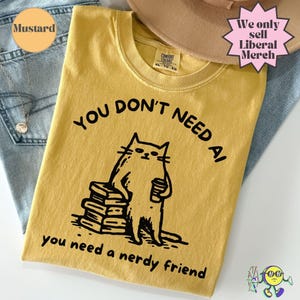 Może przedstawiać: Musztardowy t-shirt z czarną grafiką przedstawiającą kota stojącego obok stosu książek i trzymającego kubek. Napis głosi: "YOU DON'T NEED AI you need a nerdy friend." Koszulka ma okrągły dekolt i metkę marki Comfort Colors.